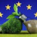 Grafika przedstawia zieloną żarówkę z trawy i żarówkę wypełnioną glebą na tle flagi Unii Europejskiej, co nawiązuje do tematu artykułu o Net Zero Industry Act